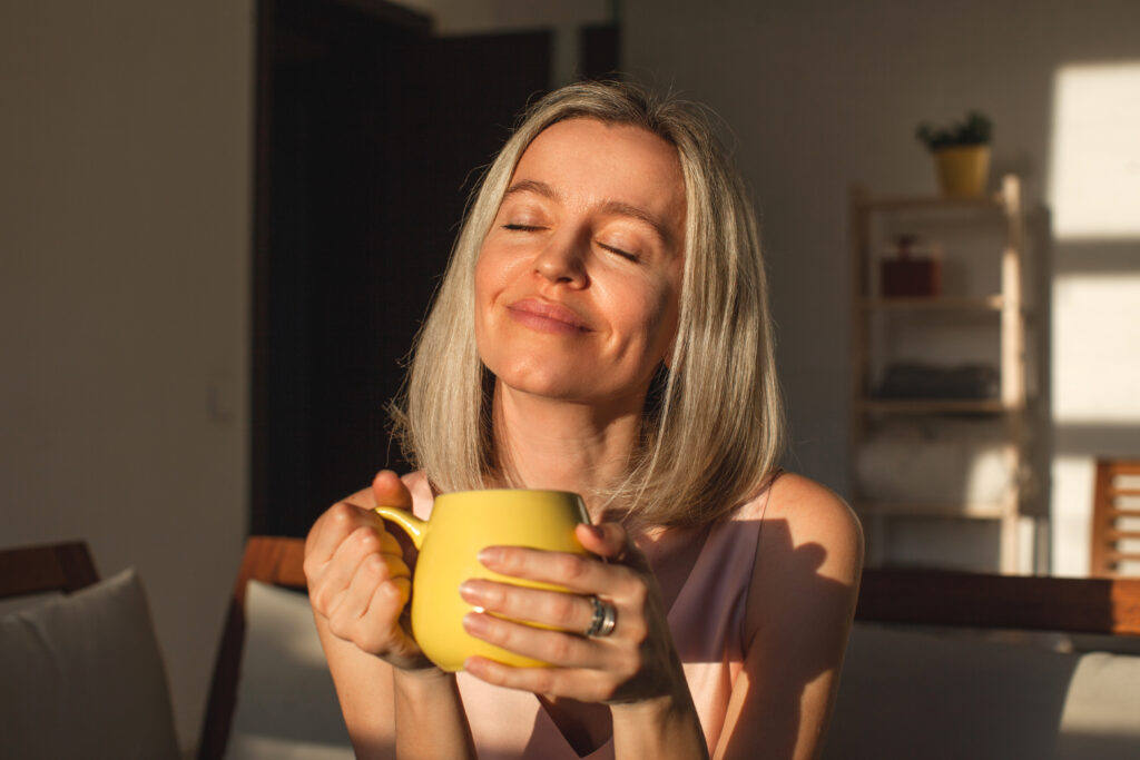 Mujer de mediana edad feliz sentada en un sofá con una taza de té con los ojos cerrados y una expresión de tranquilidad 